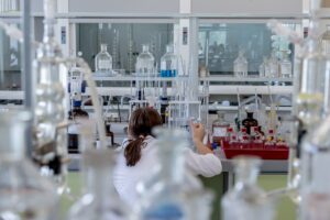 A person in a white lab coat works in a laboratory filled with various glassware and chemicals. Shelves lined with bottles and equipment are seen, as the person focuses on a task at the workbench.