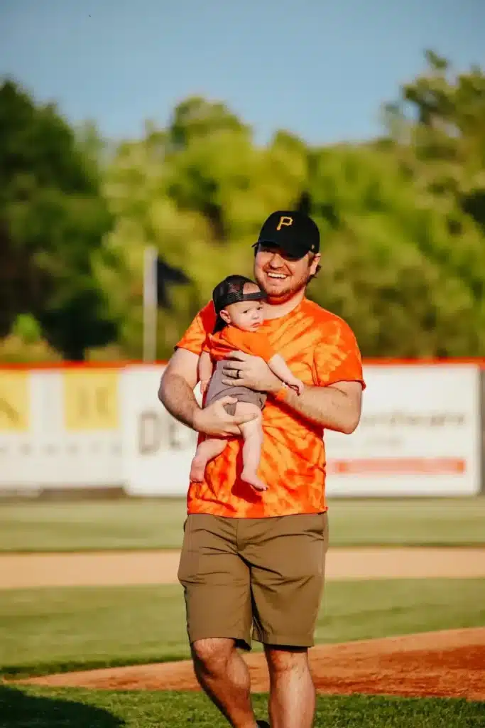 A man in an orange tie-dye shirt and baseball cap walks on a baseball field while holding a baby. Both are wearing caps, and the sunny setting shows trees and a colorful scoreboard in the background.