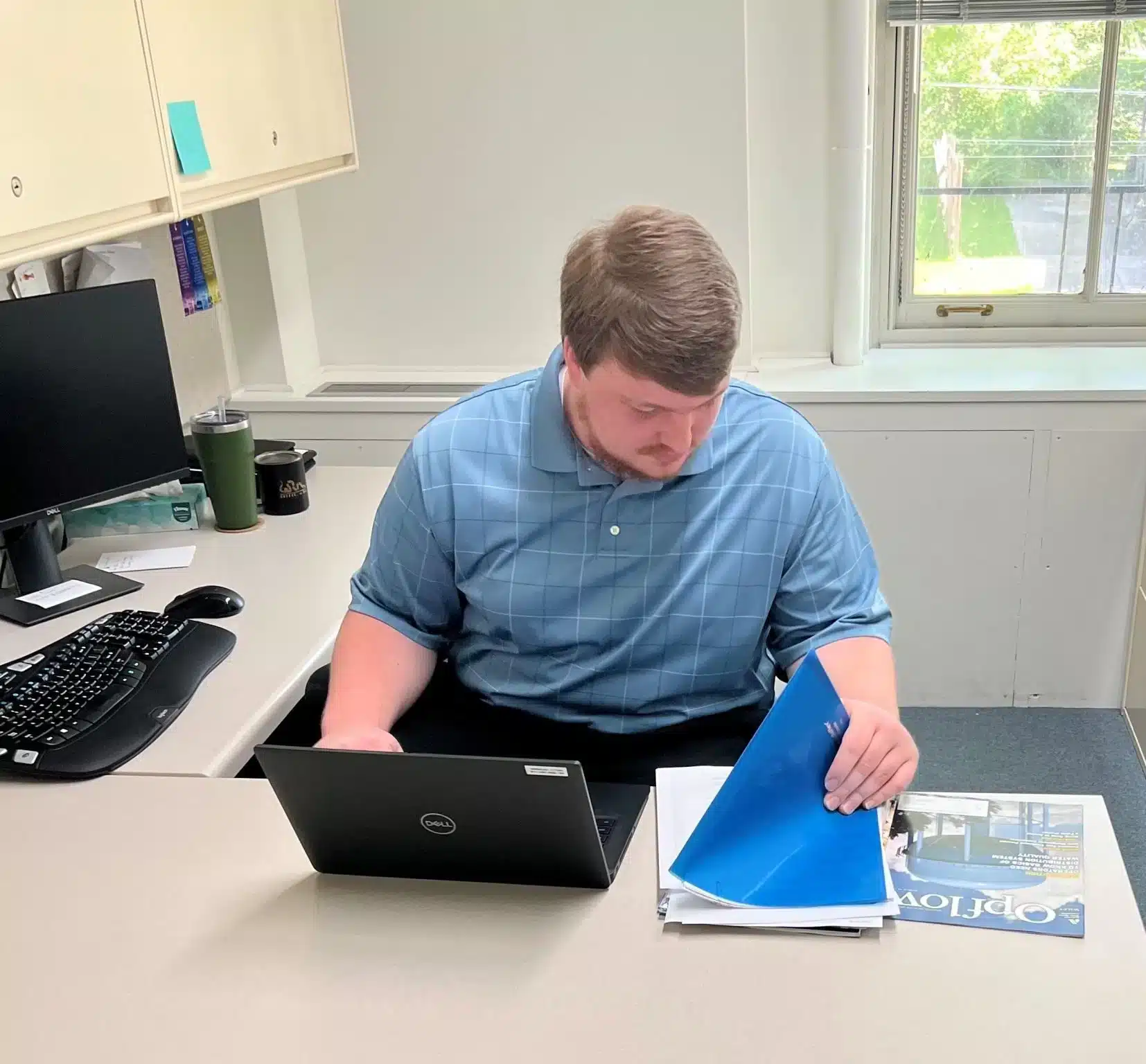 A person with short hair sits at a desk working on a laptop. They are wearing a light blue polo shirt and holding a blue folder. The desk has a keyboard, monitor, closed magazine, and a tumbler. Natural light comes through a window in the background.