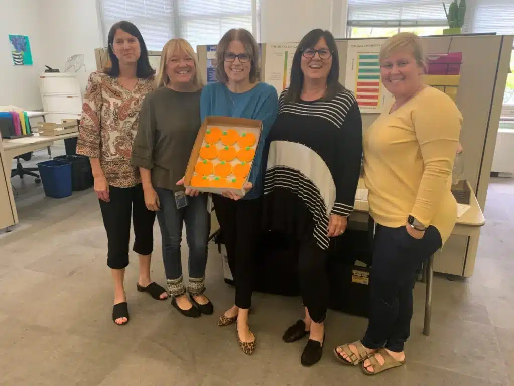 Five women stand in an office, smiling. The woman in the middle holds a tray with orange frosted cupcakes. Behind them are desks with organized papers and colorful charts on the walls.