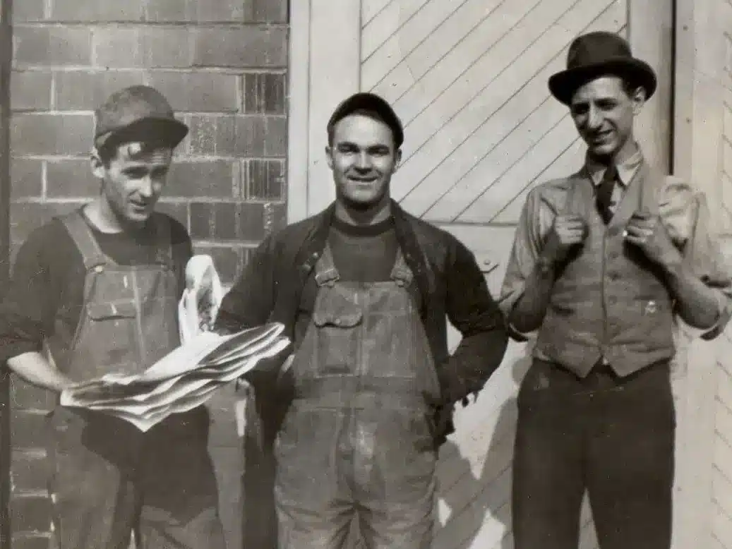 Three men pose in front of a brick building. The first holds a newspaper, wearing overalls and a cap. The second, in the center, smiles, also in overalls. The third wears a vest, tie, and hat, holding his suspenders. They convey a casual 20th-century vibe.