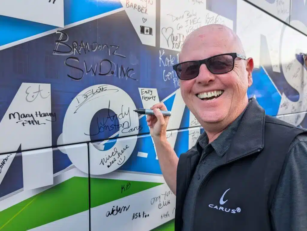 A man wearing sunglasses and a dark vest is smiling while signing a white and blue wall covered with various other signatures and messages. He holds a black marker in his right hand.