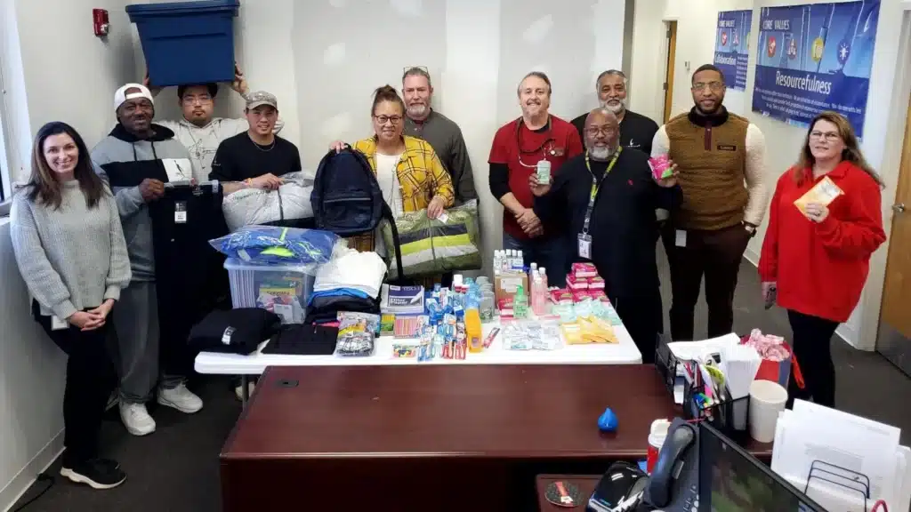 A group of thirteen people stand in a room, holding various items like toiletries and a backpack. A table in front of them displays personal care products and a blanket. They are smiling and appear to be part of a community or charity event.