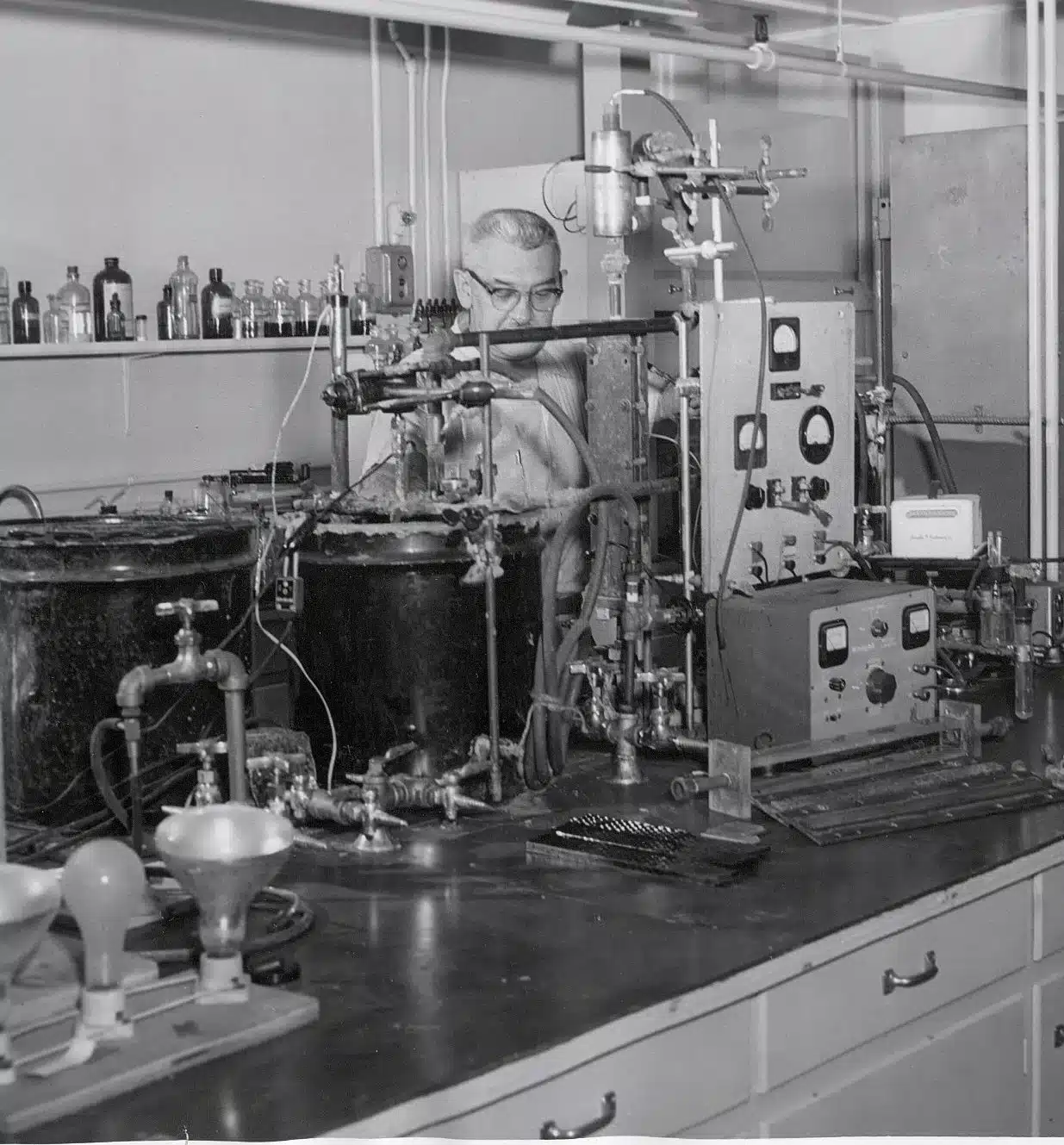 A man in a laboratory filled with scientific equipment and instruments. He is surrounded by large machines, dials, pipes, and bottles. The scene reflects a busy and intricate scientific workspace.