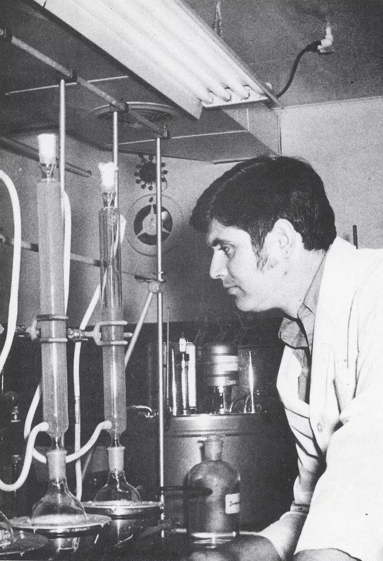 A scientist in a lab coat examines glass equipment with tubes and flasks in a laboratory setting. Shelves and various lab apparatus are visible in the background.