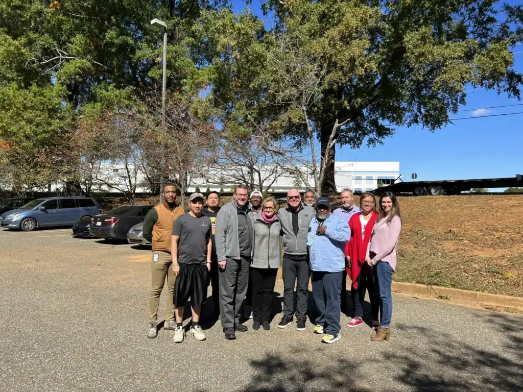 A group of ten people standing outside on a sunny day. They are posing for a photo in front of a parking lot with cars and trees. Some are wearing casual clothing and jackets. The sky is clear and blue.