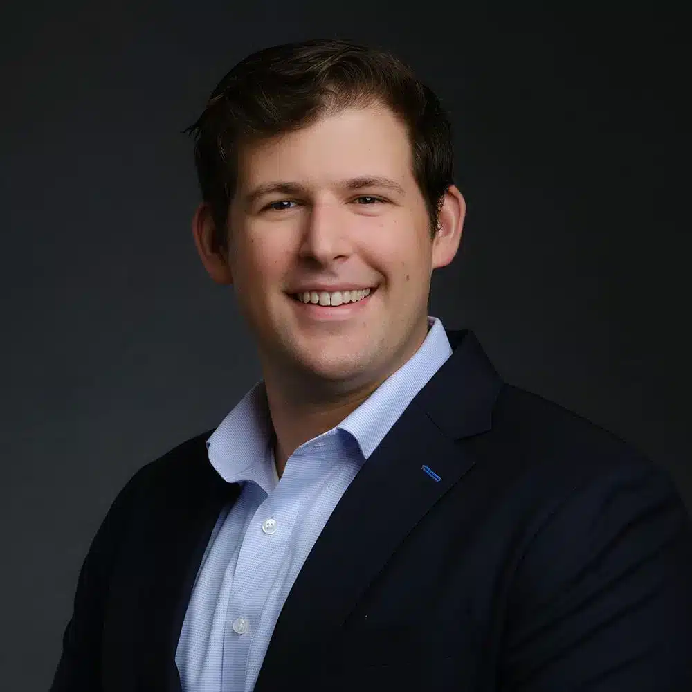 A smiling man in a dark suit jacket and light blue shirt on a dark background.