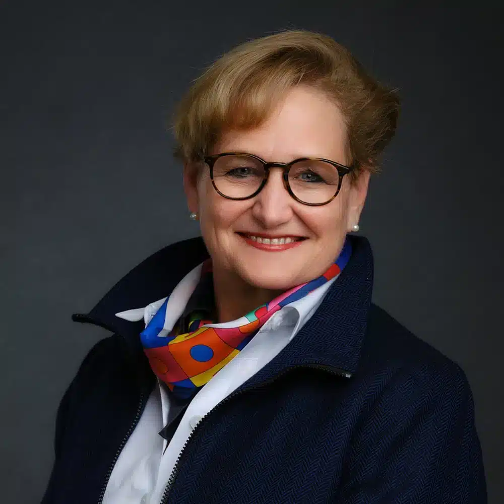 A woman wearing glasses and a bright, multicolored scarf, smiles against a dark background. They are dressed in a dark jacket over a white shirt.