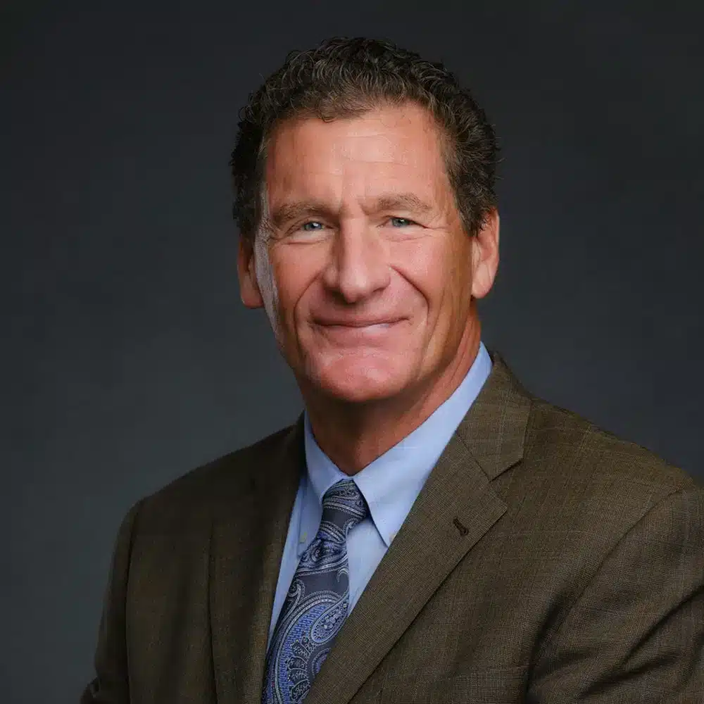 A man in a brown suit and blue patterned tie smiles against a dark background.