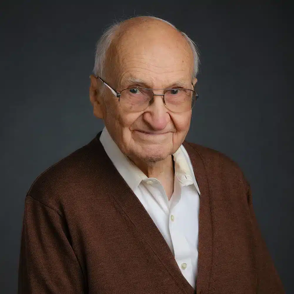 A smiling man with glasses is wearing a brown cardigan over a white shirt. The background is a neutral dark color.