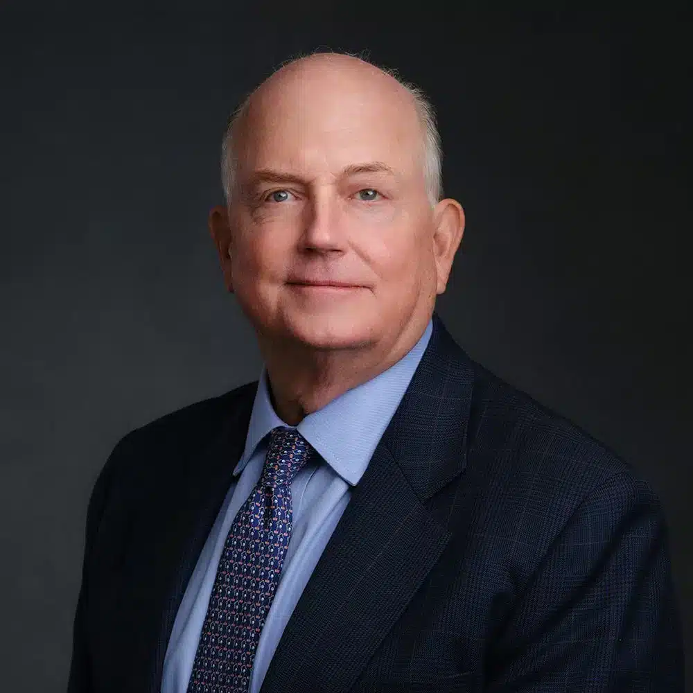 A man wearing a dark suit, blue shirt, and patterned tie, poses against a dark background.