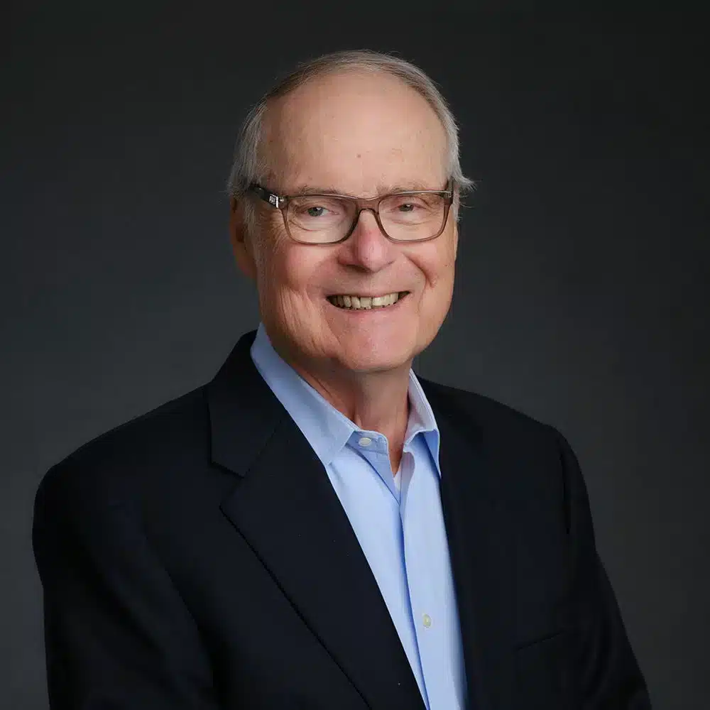 A man with glasses smiles against a dark background. He wears a light blue shirt and a dark blazer.