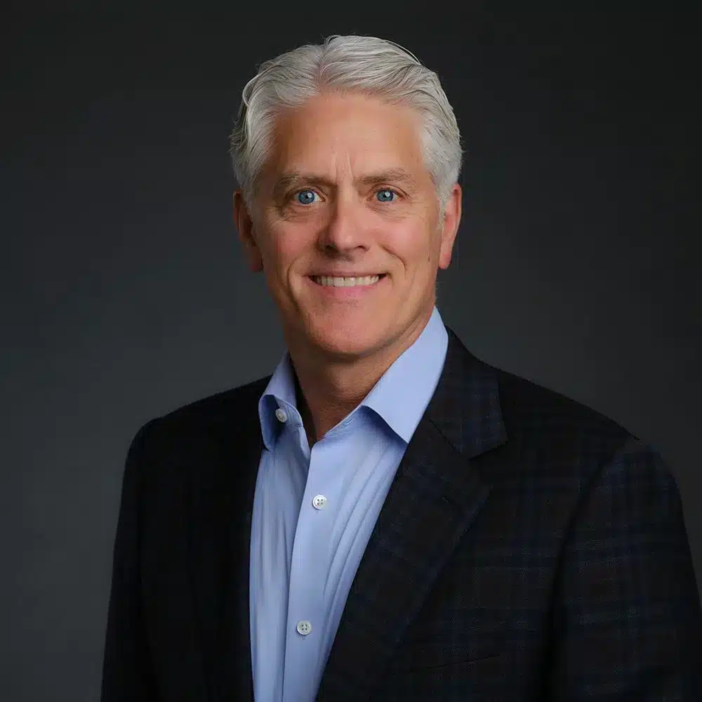 A man smiles, wearing a blue shirt and dark suit jacket, against a dark background.