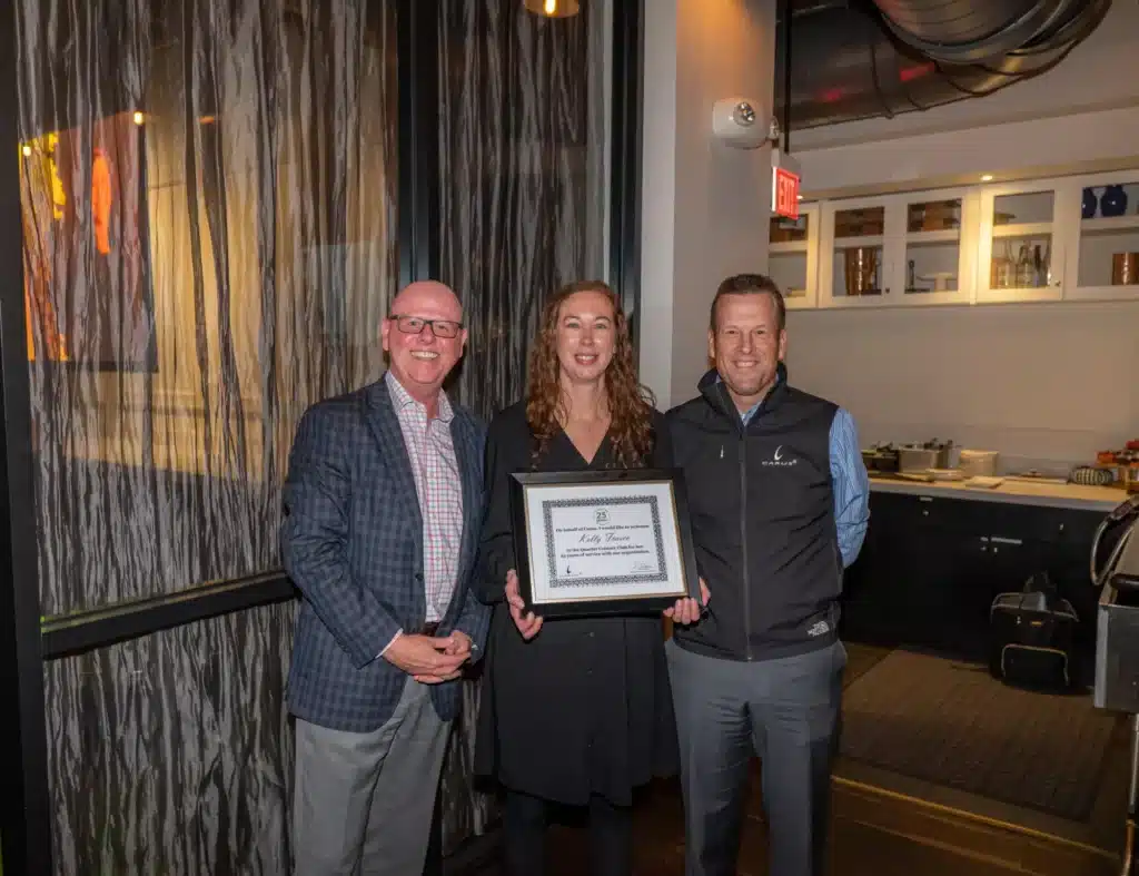 Three people in a room, the woman in the middle holding a framed certificate while smiling. Two men stand on either side of her, all dressed in business attire. The room has a glass wall, shelves, and various items in the background.