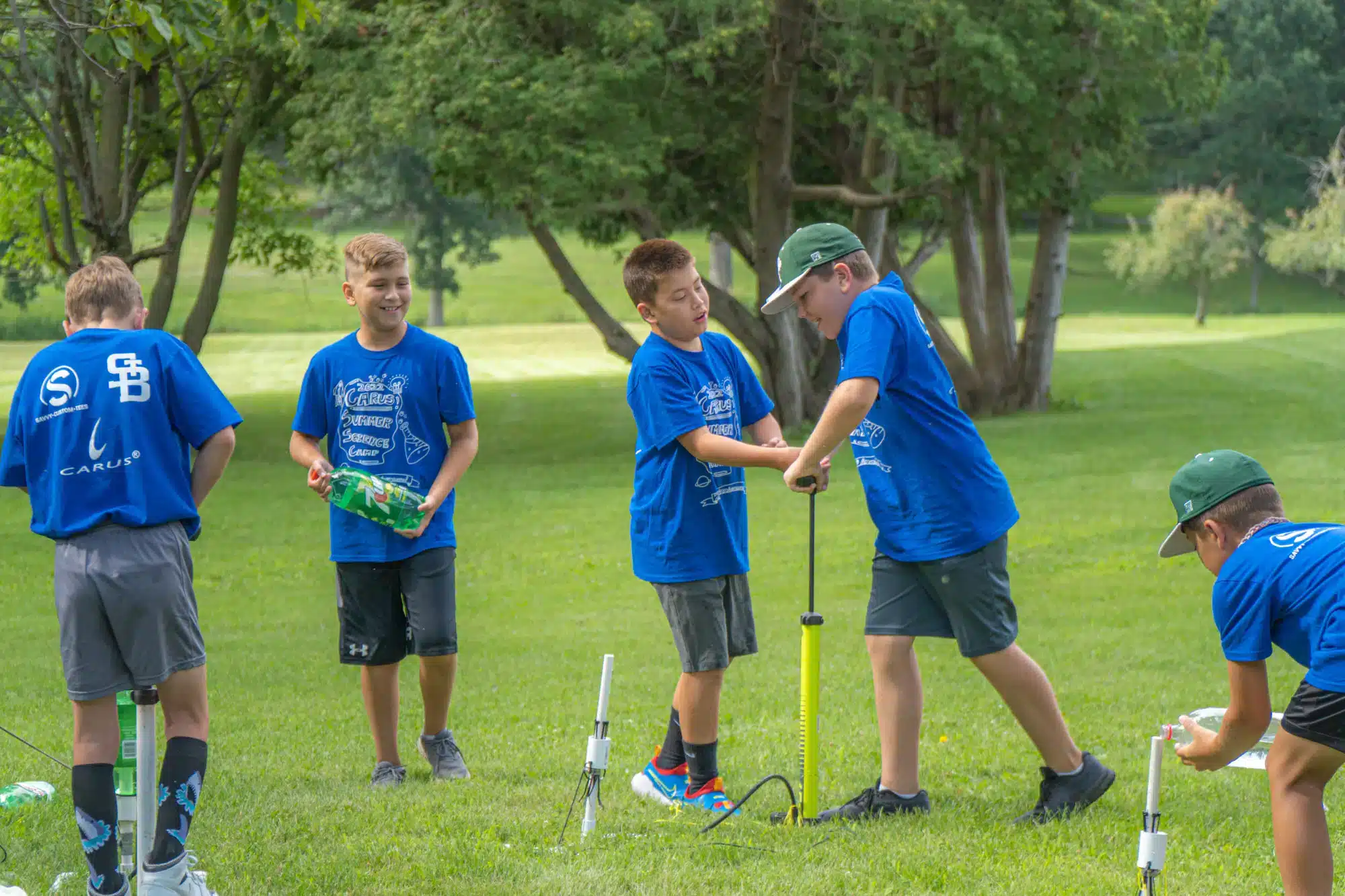 Boys in blue shirts and shorts are launching model rockets in a park. One uses a pump while others prepare the rockets. They're surrounded by green grass and trees on a sunny day.