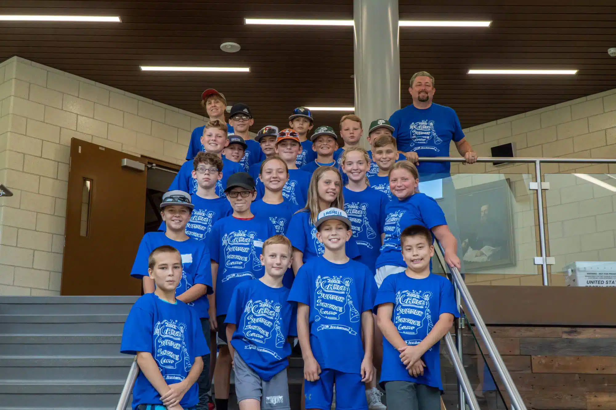 A group of children and adults wearing matching blue T-shirts pose on indoor stairs. They smile at the camera, and a man stands on a landing beside them. The background features a brick wall and modern lighting.