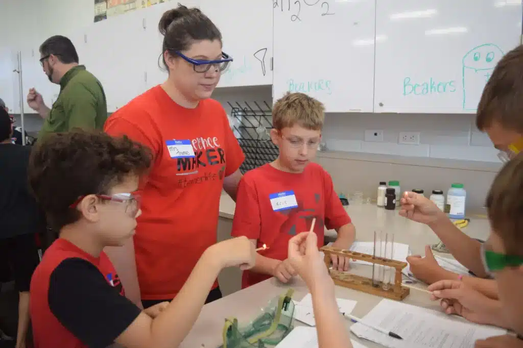 A teacher in a red shirt assists three children in a science lab. They wear safety goggles and work with test tubes and a flame. The setting is bright and educational, with supplies and beakers visible in the background.