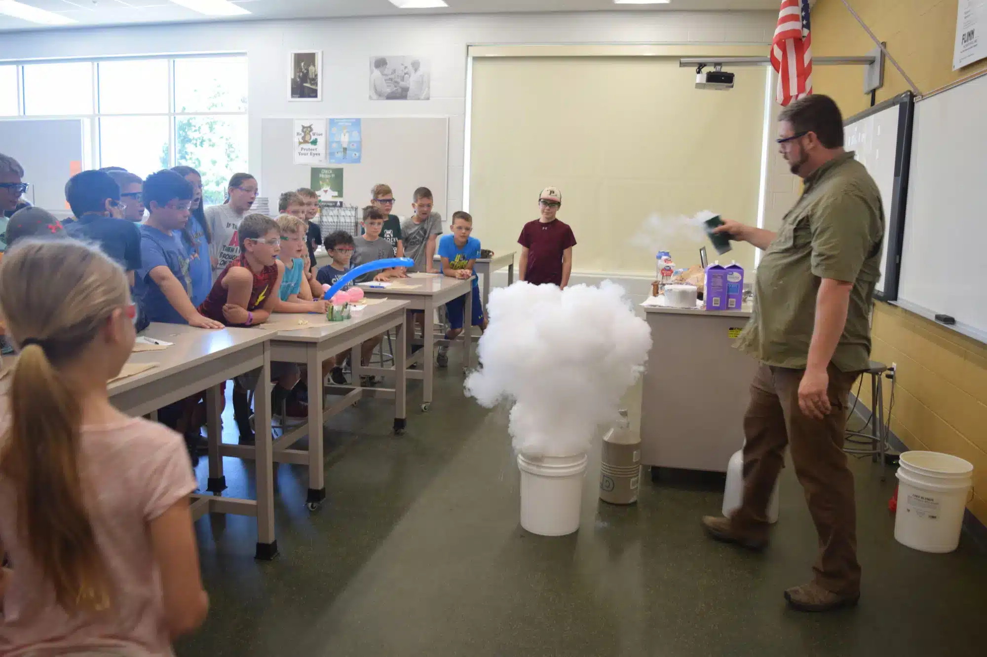 A teacher conducts a science experiment with liquid nitrogen in a classroom. Onlookers, including children, watch as a cloud of vapor rises from a bucket. The room has desks, a whiteboard, and posters, and an American flag is visible in the corner.