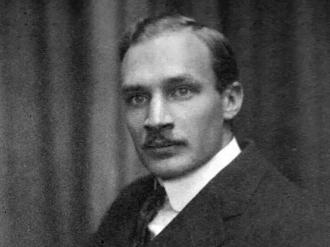 Black and white portrait of a man with a combed-back hairstyle and a mustache, wearing a dark suit and tie. He gazes directly at the camera with a neutral expression. The background is a softly blurred curtain.