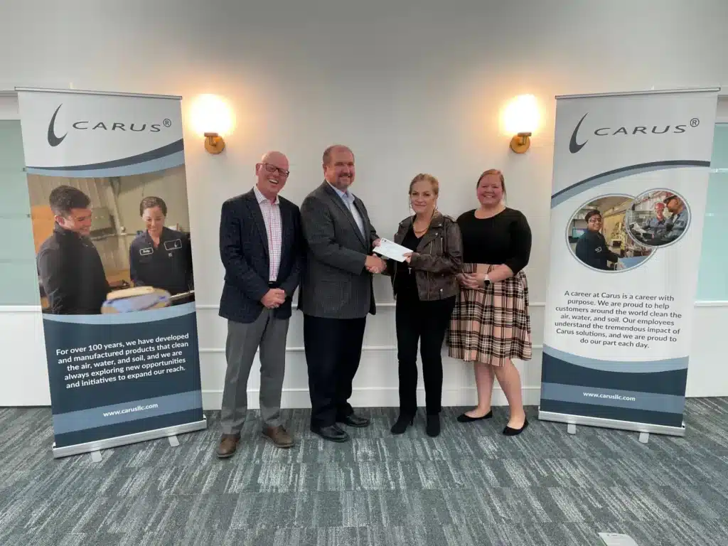 Four people stand indoors, smiling. Two men wear suits, while two women are in casual business attire. They are between two banners with the Carus logo. One man hands a woman a document or card.