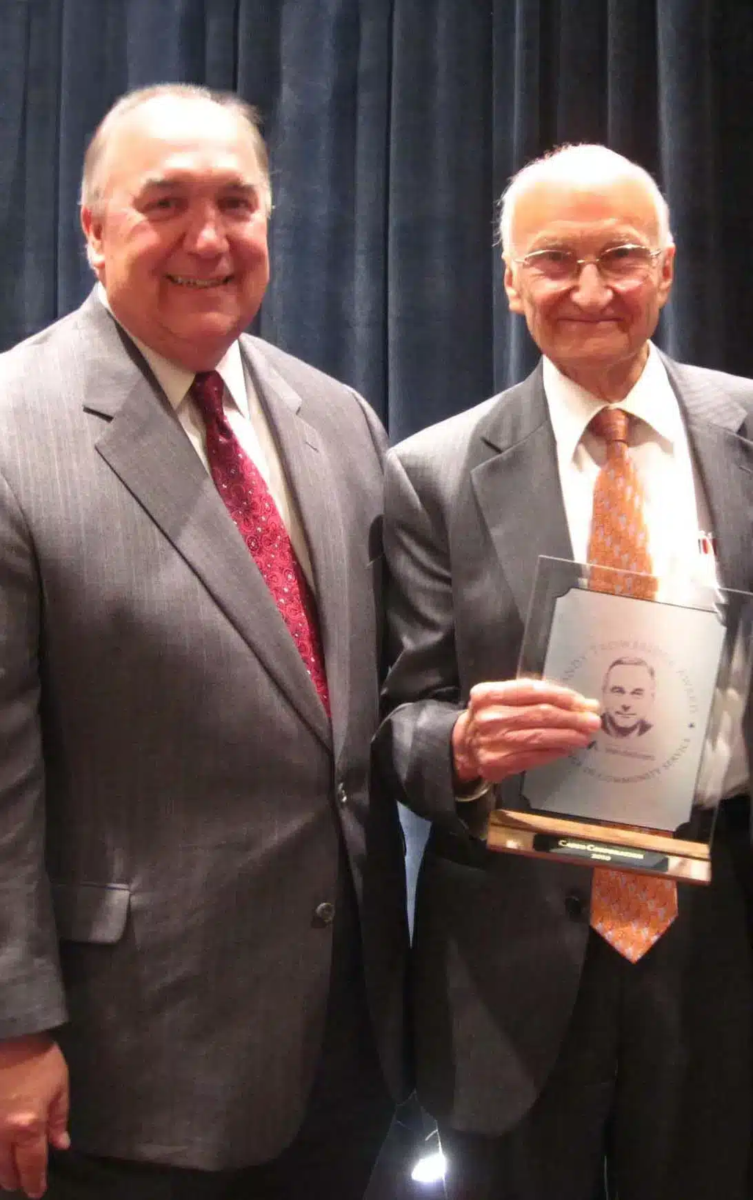 Two men in suits stand together smiling. The man on the right holds a framed award with a portrait on it. Both are in front of a dark curtain backdrop.