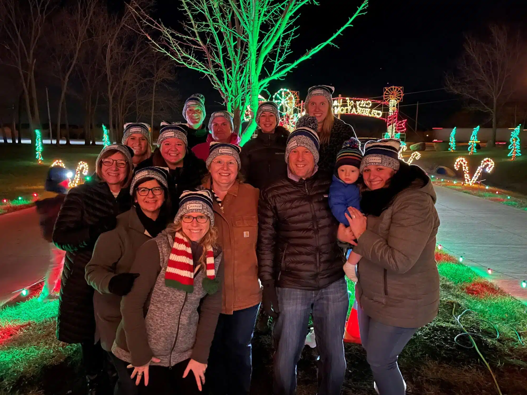 A group of people dressed warmly, wearing matching beanies, pose together outdoors at night. Behind them are festive holiday lights, including trees and candy canes illuminated in red and green. A child is held by one of the adults.