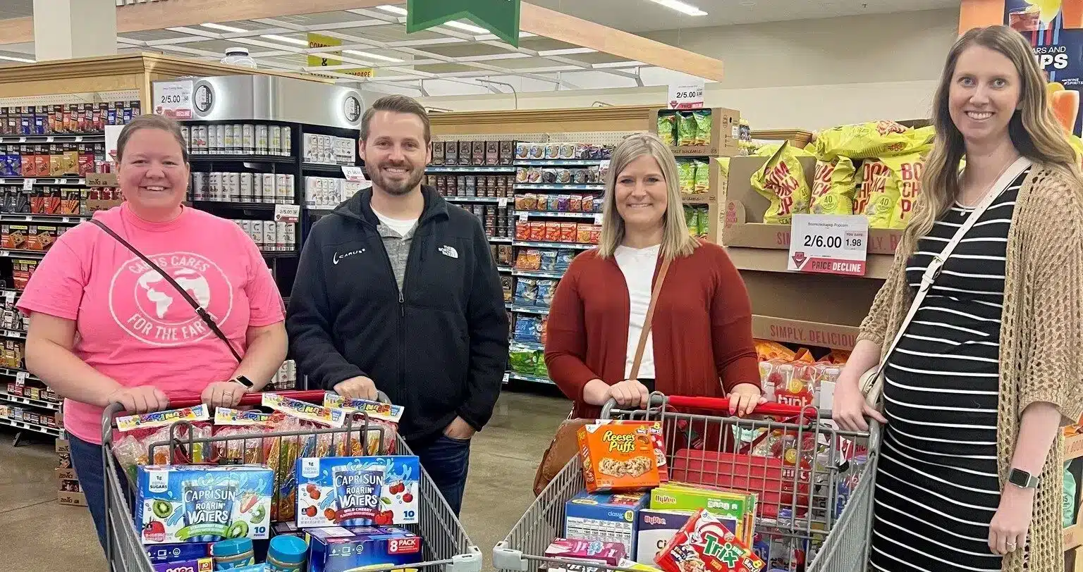 Four people are standing in a grocery store with two shopping carts full of various food items. The group appears to be smiling and posing for the photo. Shelves with snacks and other products are visible in the background.