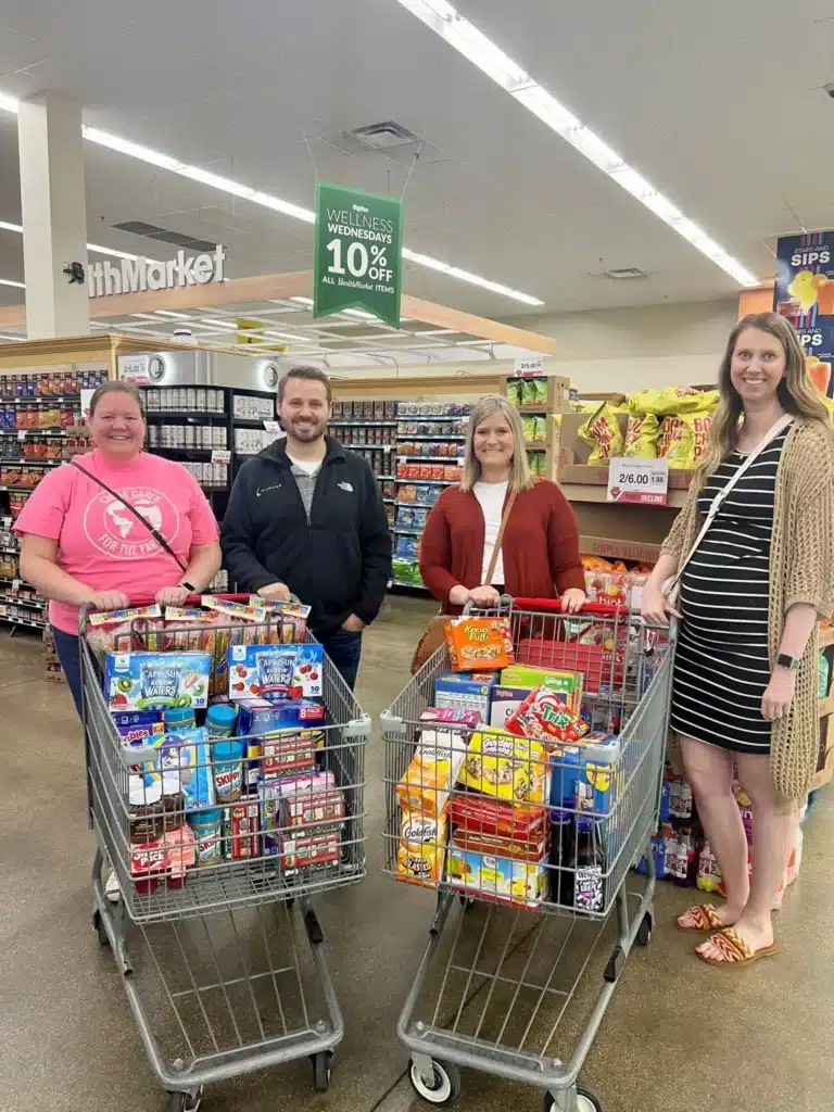 Four people stand in a supermarket aisle with two shopping carts filled with various groceries, including snacks. There's a sign above them announcing a Wednesday discount. Shelves stocked with products line the background.