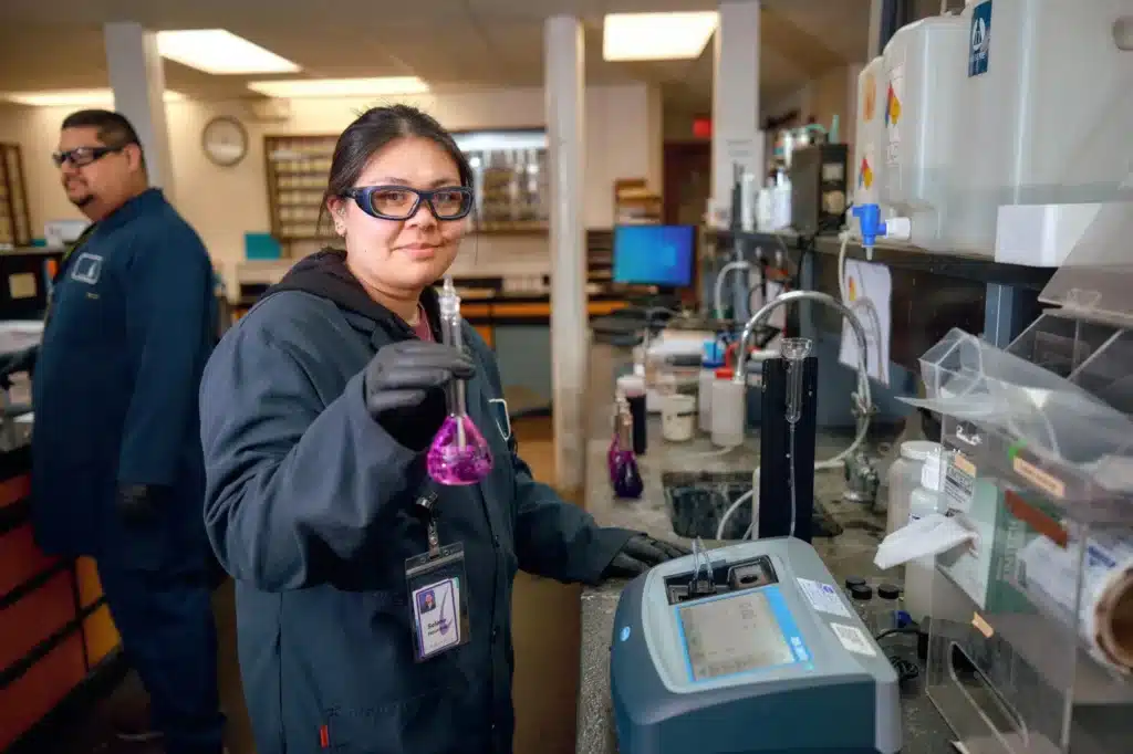 A person wearing safety goggles and a lab coat holds a flask with purple liquid in a laboratory. Another person in similar attire stands in the background. The lab features various equipment and chemical containers.