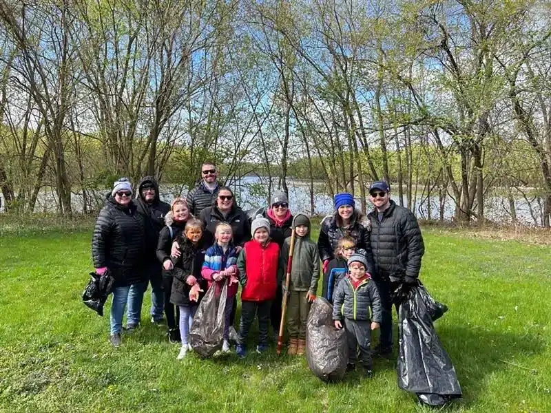 A group of adults and children stand on grass, smiling and holding large garbage bags. They are dressed in warm clothing, surrounded by trees with a body of water in the background. The scene suggests a community cleanup activity.