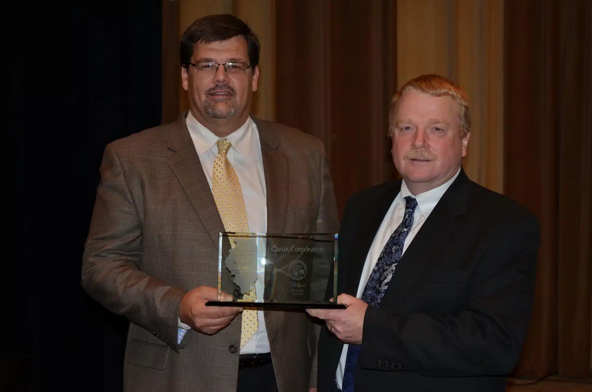 Two men in suits holding a glass award. One has a gray suit with a yellow tie, the other a dark suit with a patterned tie. They stand in front of a dark drape and wooden paneling, looking at the camera.