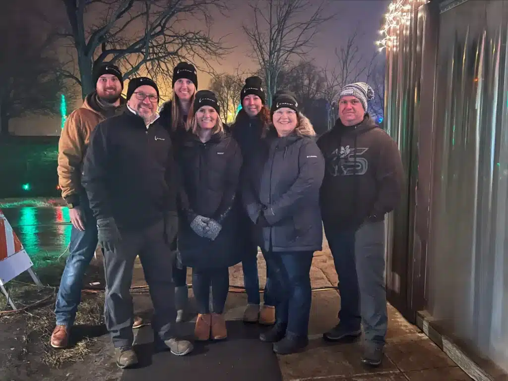 A group of seven people, wearing winter coats and hats, stand outside at night. They smile at the camera, with trees and subtle holiday lights visible in the background, creating a festive atmosphere.