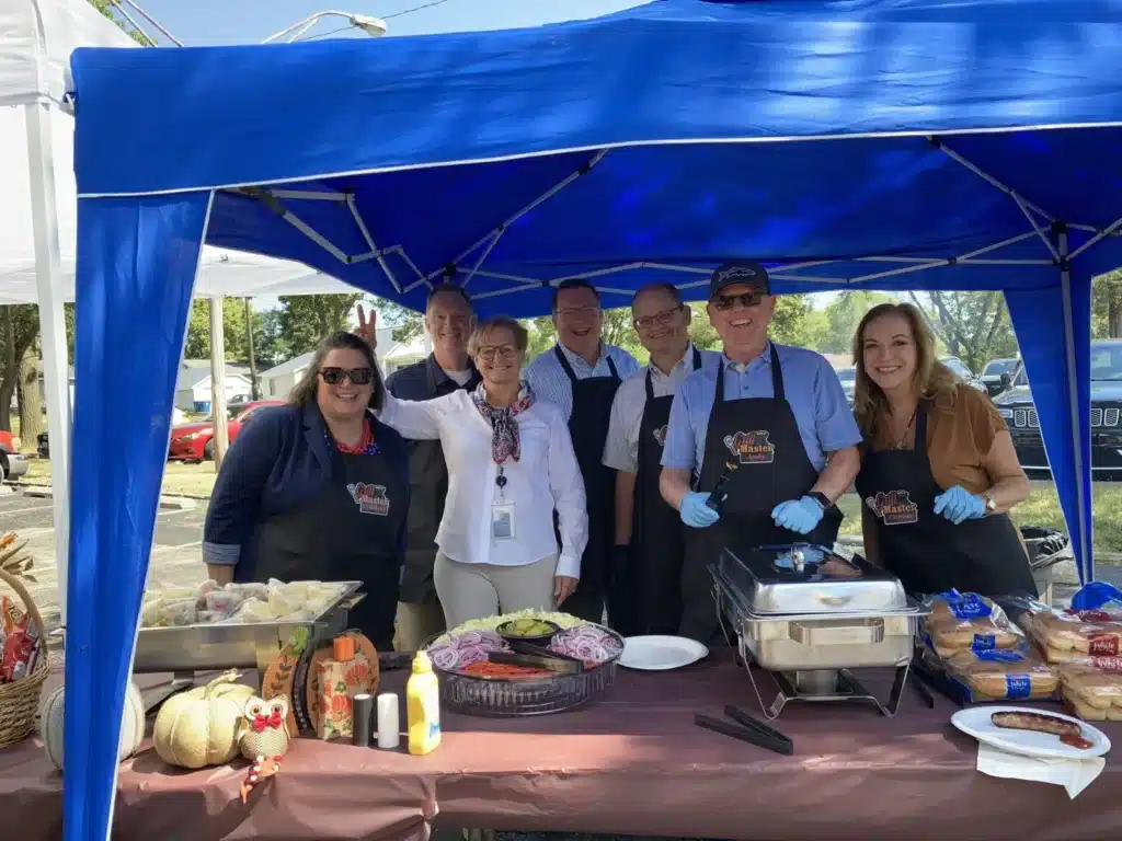 A group of seven people stands under a blue canopy at an outdoor event. They are smiling, wearing aprons, and serving food from chafing dishes. A table with food items, decorations, and a basket is in front of them.