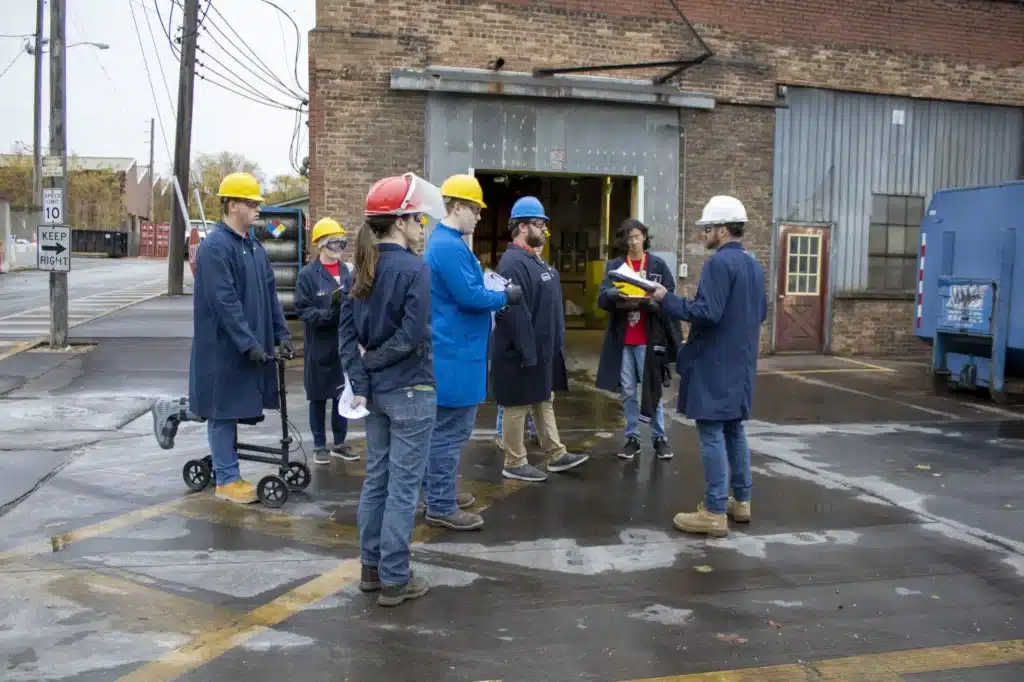 A group of people wearing hard hats and safety jackets stand in a circle on a wet pavement outside an industrial building. Some are holding notepads, and one is on a scooter. A large blue container is visible in the background.