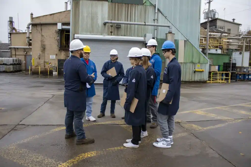 A group of seven people wearing hard hats and protective coats are standing in a circle in an industrial setting. They appear to be discussing something. Some are holding clipboards. The background shows industrial buildings and equipment.