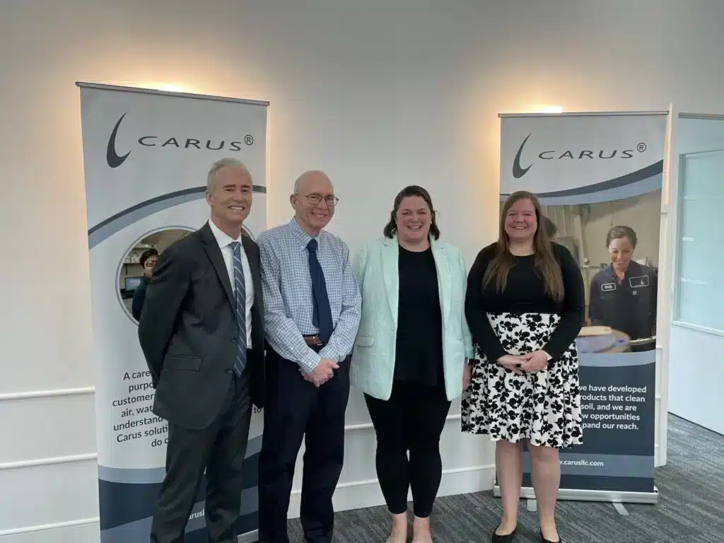 Four people standing and smiling in front of two Carus promotional banners in an office setting. Three are wearing business attire, and one is in a floral skirt and black top.