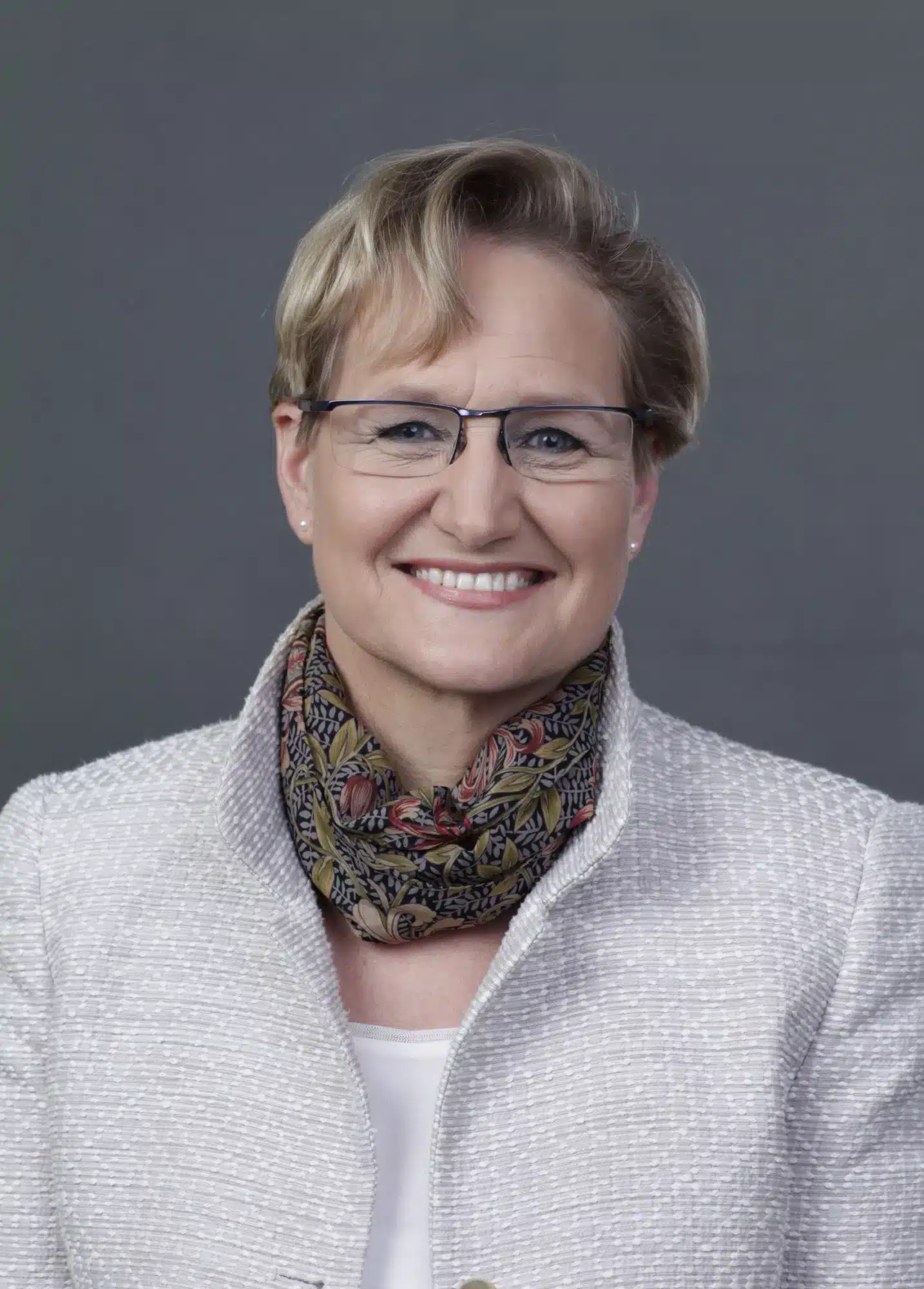 A woman with glasses smiles while wearing a light gray blazer and a patterned scarf against a plain gray background.