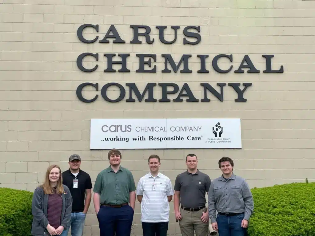 Six people stand in front of the Carus Chemical Company building. They are smiling and dressed in casual to business-casual attire. A sign behind them reads, "working with Responsible Care." Bushes are visible at the bottom.