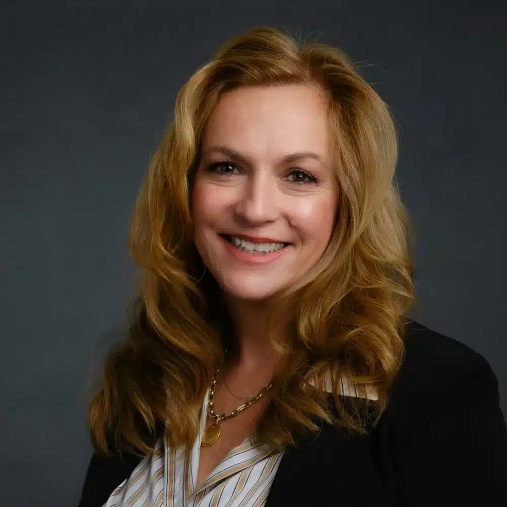 Woman with long, wavy hair smiling, wearing a dark blazer and striped blouse, with a necklace. Neutral gray background.