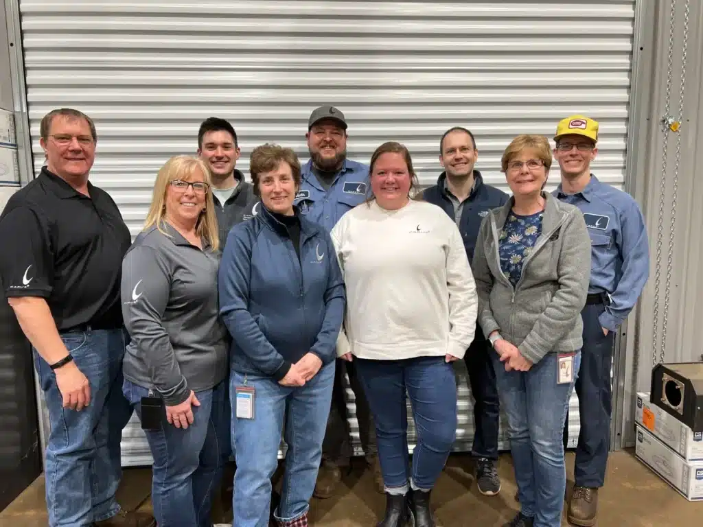 A group of nine people, five men and four women, stand smiling in front of a corrugated metal wall. They are wearing casual work attire, including jeans, jackets, and polos. The setting appears to be industrial or workplace-related.