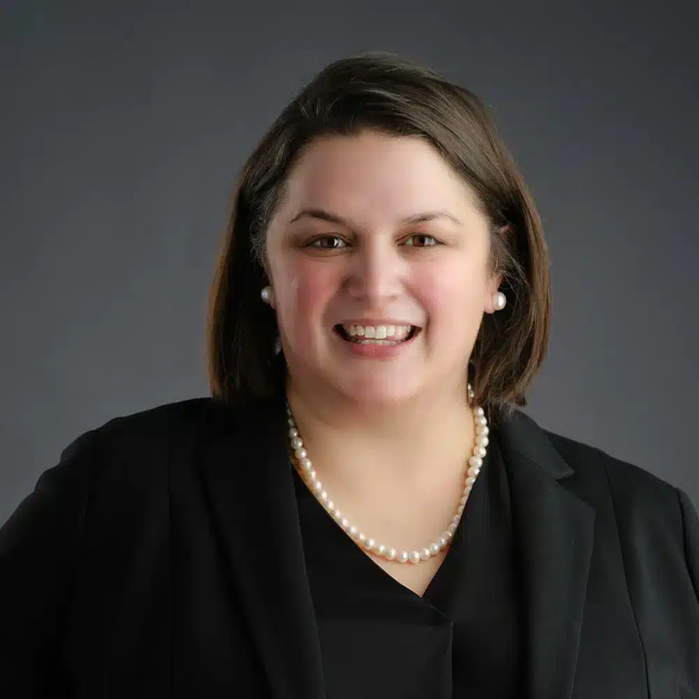 A smiling woman, wearing a black blazer, black top, and a pearl necklace. They have shoulder-length brown hair and are set against a plain dark background.