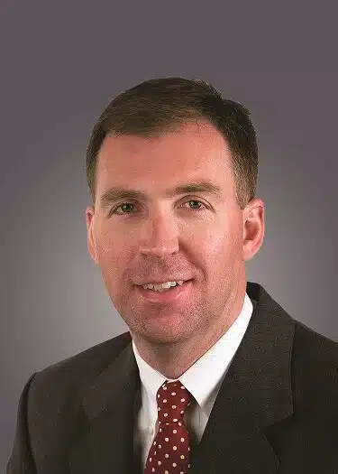 A man in a dark suit and polka dot tie is smiling against a plain gray background.