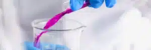 A person wearing blue gloves carefully pours a vibrant pink liquid from a test tube into a clear beaker. The laboratory setting is blurred in the background, emphasizing the careful handling of the colorful substance.