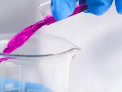 A person wearing blue gloves carefully pours a vibrant pink liquid from a test tube into a clear beaker. The laboratory setting is blurred in the background, emphasizing the careful handling of the colorful substance.