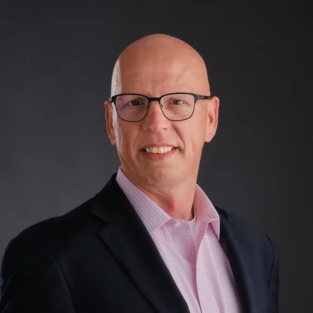 A man with glasses is smiling in a professional headshot. He is wearing a dark suit jacket over a light pink shirt. The background is a plain dark grey.