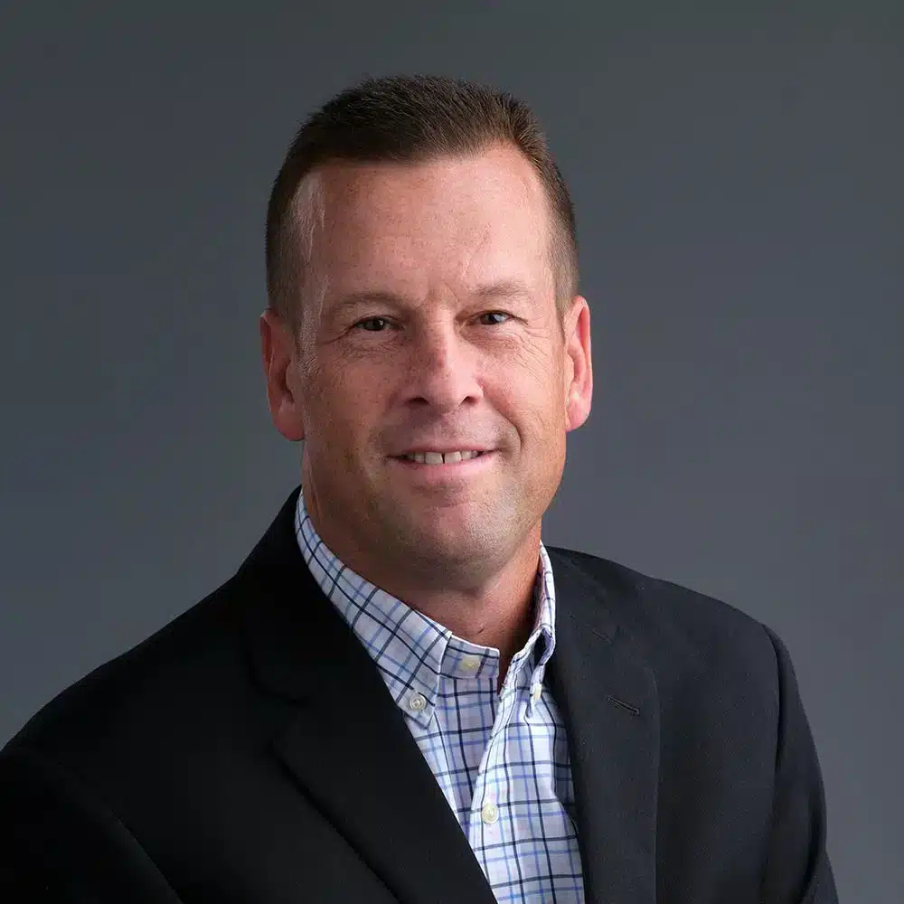 A professional portrait of a person with short hair, wearing a dark suit jacket over a checkered shirt. The background is solid gray.