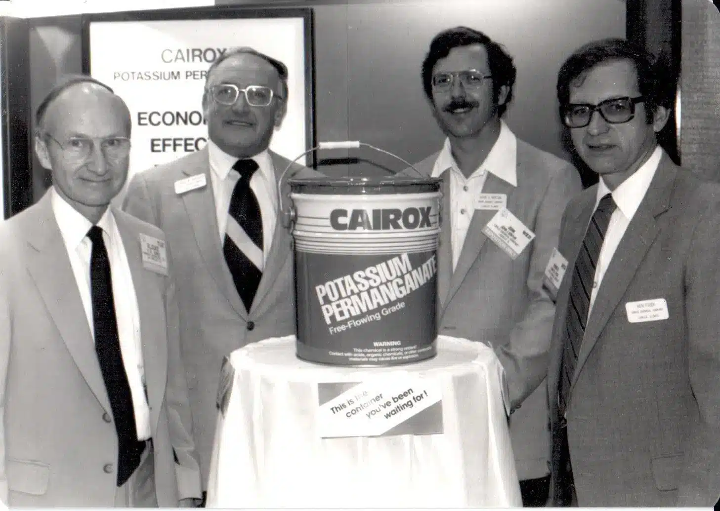 Four men in suits stand around a display featuring a large container labeled "Cairox Potassium Permanganate." They are smiling and wearing name tags. There is a sign in the background about the product's economic effectiveness.