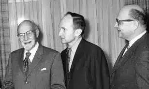 Three men in suits stand together, smiling and engaged in conversation. The setting appears formal with draped curtains in the background, suggesting a gathering or event.