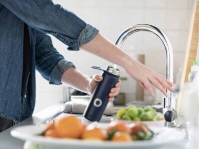 A person wearing a blue shirt fills a blue reusable water bottle at a kitchen sink. The countertop holds a plate of oranges and various vegetables. The room is bright and airy.