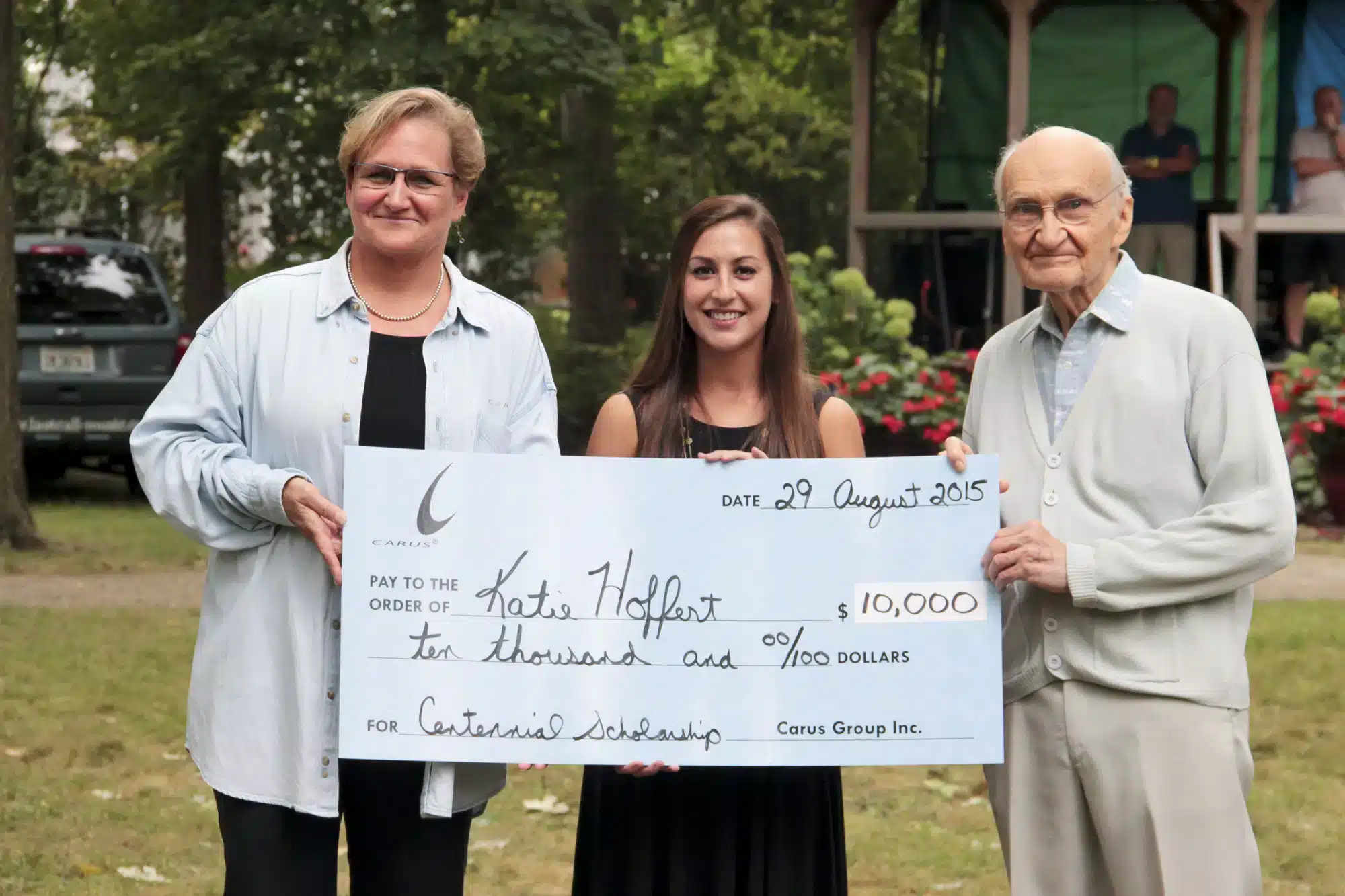 Three people standing outdoors hold a large check for $10,000, made out to Kate Holfert for an academic scholarship dated August 29, 2015. They appear happy and proud, with a stage and trees in the background.